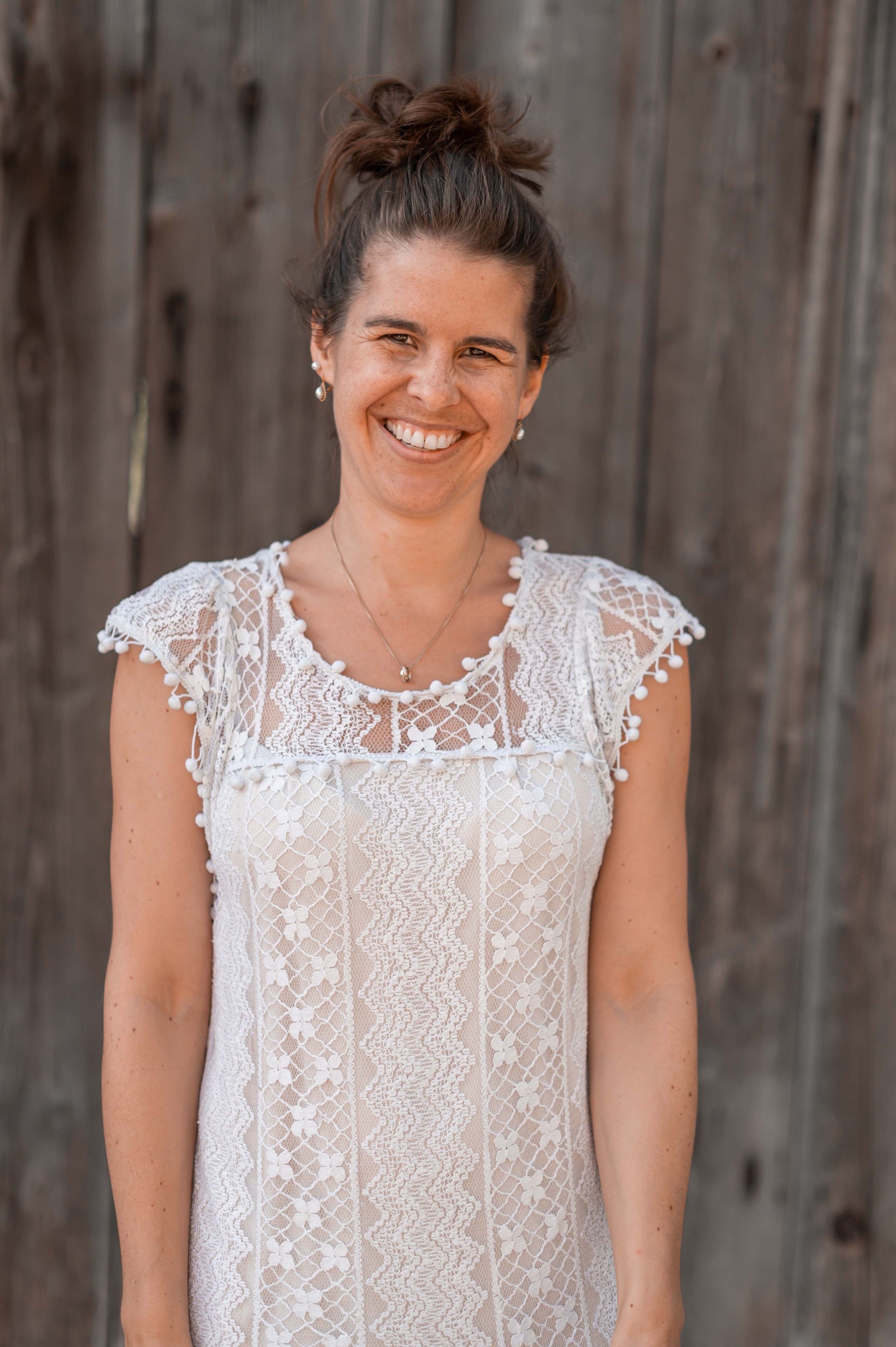 Lächelnde weibliche Person mit langen braunen Haaren, welche zu einem Dutt zusammengebunden sind. Sie trägt Ohrschmuck, eine goldige Halskette und ein weisses Oberteil. Ihr Blick strahlt und sie zeigt mit ihrem breiten Lächeln ihre gesunden Zähne.